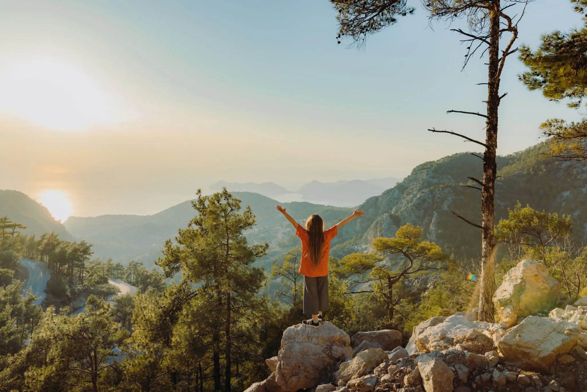 Person in orange jacket enjoying mountain sunrise with arms raised.